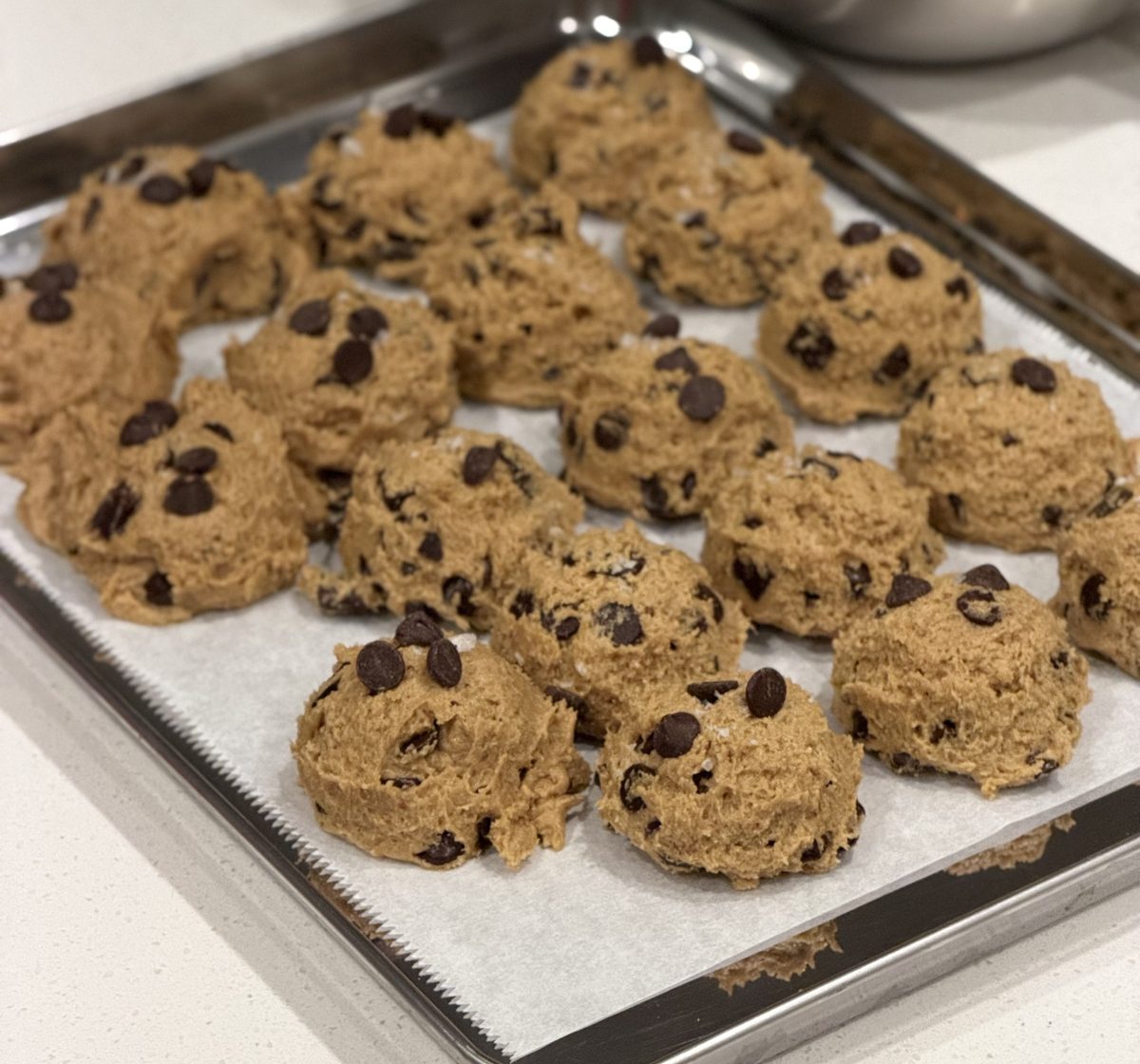 Eighteen scooped balls of cookie dough studded with chocolate chips on a parchment-lined sheet pan